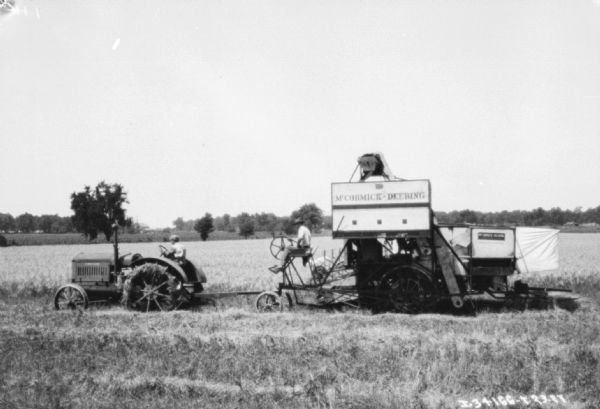 Left side profile view of a man driving a tractor pulling a man sitting on a harvester thresher in a field.