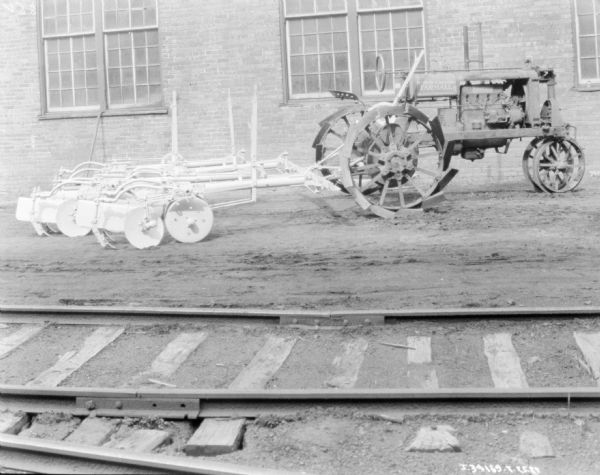 View across railroad tracks towards a tractor, on the right, hitched to a disk plow, on the left. In the background is a brick factory building.