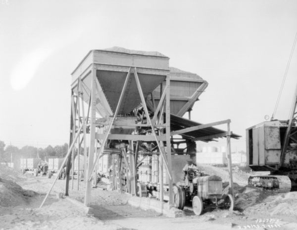Men driving tractors pulling trailers, which are being loaded from a hopper at a granite quarry or mine. On the far right is a steam shovel.