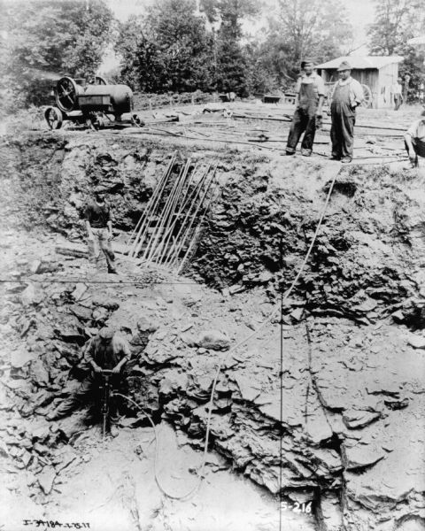 A man is working down in a trench with a tool attached by a hose to machinery parked near the top of the trench. Other men are standing above the trench watching.