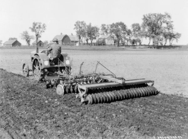 Three-quarter view from left rear of a man driving a 10-20 tractors pulling diskharrow and culti-packer. Farm buildings are in the background among trees.