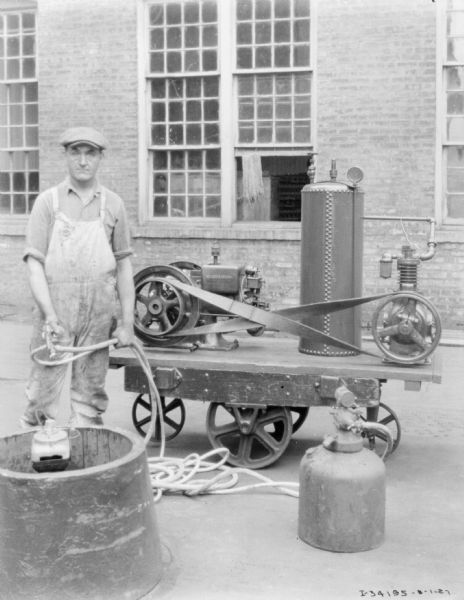 A man is standing outdoors near an engine. There is a brick factory building in the background.
