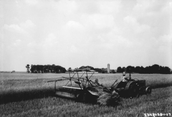 View across field towards a man driving a tractor pulling a binder. There are farm buildings in the background among trees.