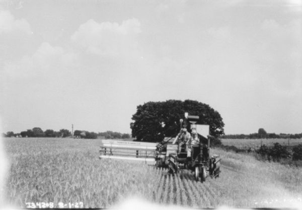 View looking down partially harvested field towards a man driving a tractor. Behind him a man is sitting on the front of the harvester thresher.