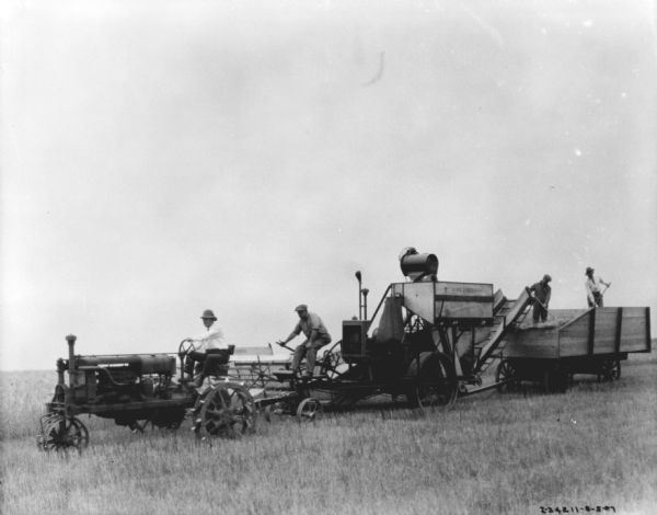 View across field towards four men working with a harvester-thresher. The man on the left is driving a tractor, another man is sitting on the front of the harvester-thresher, and two men are standing in a wagon on the right in a pile of hay.