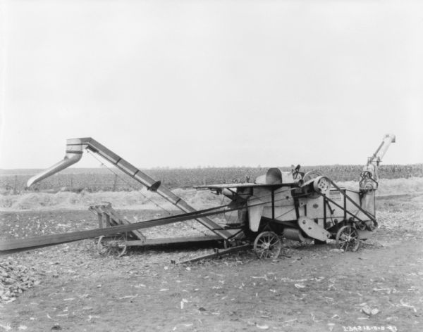 A harvester-thresher, shredder is being belt-driven in a field. In the background are piles of hay, and a fence and a field are in the far background.