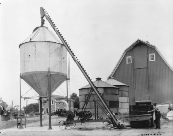 A man is standing on the right near an ensilage cutter and truck. The silo is being filled. Two storage tanks and a barn are in the background.