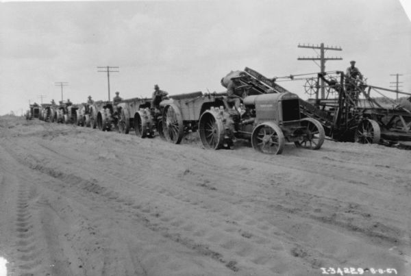 View towards a long line of men sitting on industrial tractors at a road construction site. Behind the tractors a man is standing and operating a Russell Elevating Grader.