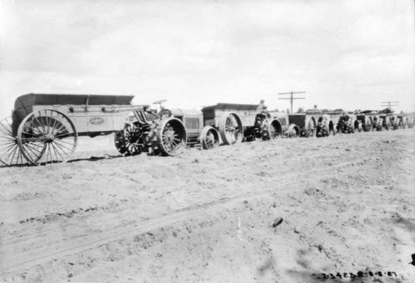 View towards a long line of men sitting on industrial tractors, each pulling a wagon, at a road construction site.