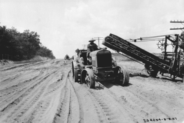 View towards a man sitting on an industrial tractor, which is pulling a wagon. On the right another man is standing and operating a Russell Elevating Grader.