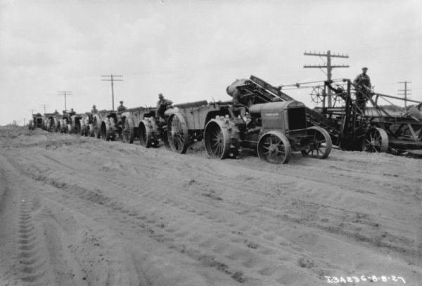 View towards a long line of men sitting on industrial tractors at a road construction site. Behind the tractors a man is standing and operating a Russell Elevating Grader.