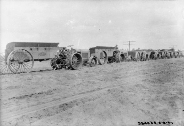 View towards a long line of industrial tractors at a road construction site. The tractors are hitched to wagons.