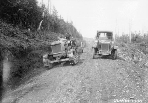 View from front looking down road towards two men grading a rough road, pulling off to side to let car pass. The man in front is driving a continuous track tractor, and the other man is standing on construction equipment pulled by the tractor to grade the road.