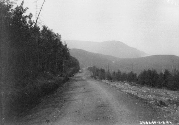 View down dirt road towards an automobile coming up the hill. Trees have recently been cleared from the side of the road, and tree-covered hills are in the distance.
