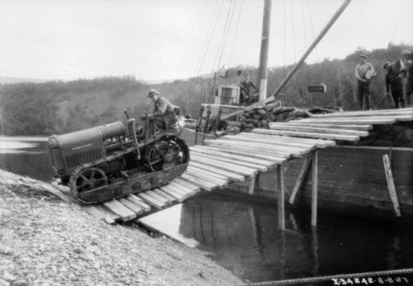 View along shoreline towards a man driving a continuous track tractor down a steep ramp over water from a boat onto the shoreline. Men are standing on the boat watching, some carrying sacks. A ridge with trees is on the opposite shoreline in the background. The tractor is for a road construction project.