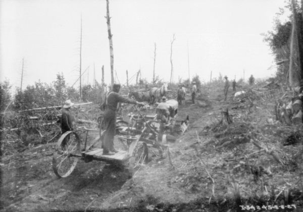 View from rear of a group of men clearing trees for road construction. A man is driving a continuous track tractor to pull a piece of construction equipment. Other men are standing near horses and downed trees in the background.