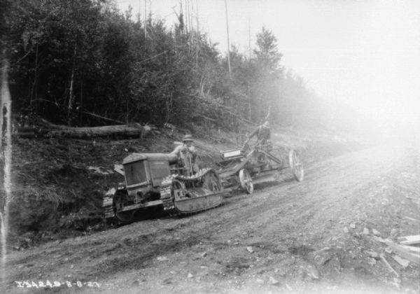 View across dirt road towards two men grading a rough road. The man in front is driving a continuous track tractor, and the other man is standing on construction equipment pulled by the tractor to grade the road. The sides of the road have cleared trees lying on the ground.