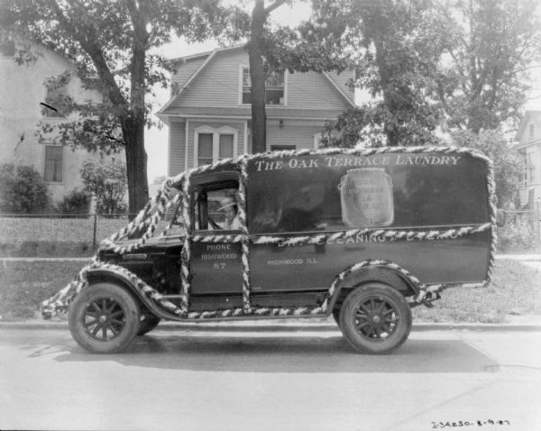 View across street towards a man sitting in the driver's seat of the Oak Terrace Laundry truck. The truck is decorated with striped swags. Houses are in the background