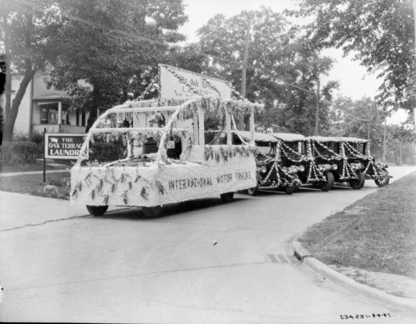 View down street towards a vehicles decorated for a parade. The side of the float on the left reads: "International Motor Trucks," and a sign on the top of the float reads: "The Oak Terrace Laundry." There is a sign for The Oak Terrace Laundry in the lawn on the left. More trucks decorated with striped swag are parked at an angle on the right.