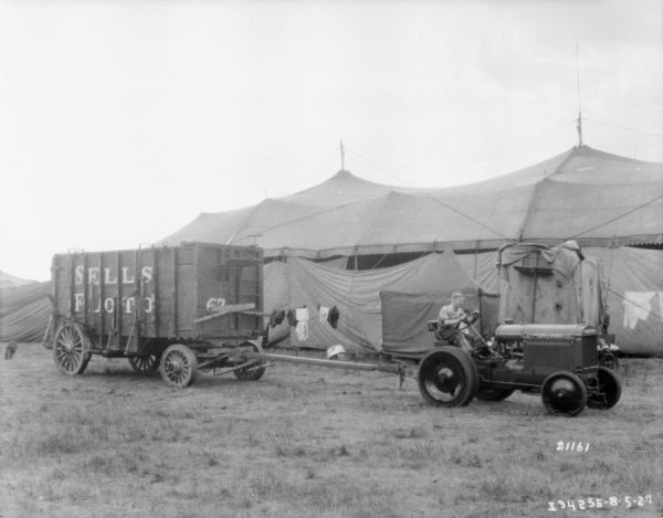 View across grounds towards a man driving a McCormick-Deering tractor pulling a Sells-Floto Circus wagon. Tents are in the background.