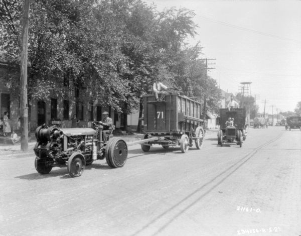 View down street towards two men driving up the street on tractors pulling Sells-Floto circus wagons. People are watching from the sidewalk across the street.