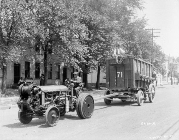 View down street towards a man driving up the street on a tractor pulling Sells-Floto circus wagons. People are watching from the sidewalk across the street.