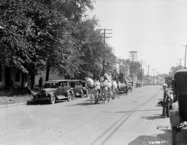 View down street towards a parade of horse-drawn circus wagons coming up the street. People are watching from the sidewalks. Automobiles are parked along the curbs on both sides of the street.