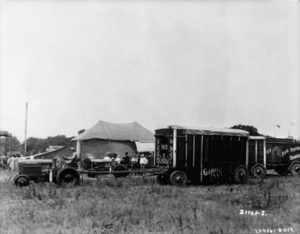 Left side view of a man driving a tractor pulling a Sells-Floto circus wagon onto a lot. Tents are in the background.