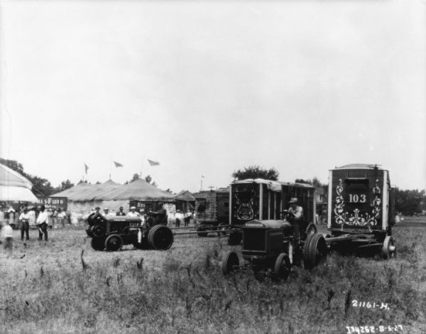 Men driving tractors are pulling Sells-Floto circus wagons onto a circus lot. Tents are in the background.
