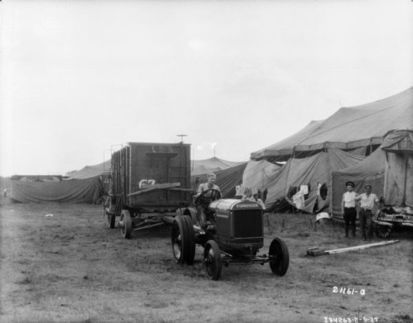 View from front of a man driving a tractor pulling a Sells-Floto circus wagon onto a circus lot. Tents are in the background.
