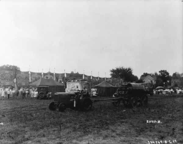 View across grounds towards a man driving a tractor pulling a water tank. A sign painted on the tank reads: "Sells-Floto Circus." People are watching in the background, near tents and circus banners.