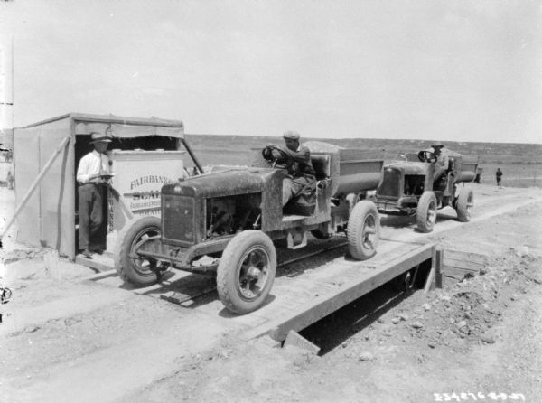 A man is sitting in the driver's seat of an open truck loaded with grain on a scale at a weigh station. A man is standing near a small building holding the scale on the left. There is another man sitting in a truck waiting to use the scale.