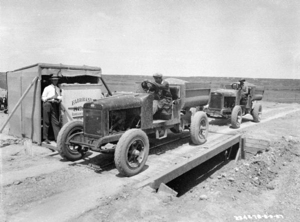 A man is sitting in the driver's seat of an open truck loaded with grain, which is sitting on a scale at a weigh station. A man is standing near a small building holding the scale on the left. There is another man sitting in a truck waiting to use the scale.