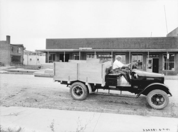 Right side view of a man sitting on the bench of an open delivery truck. There is lumber stacked in the truck bed. On the other side of the street is a storefront.