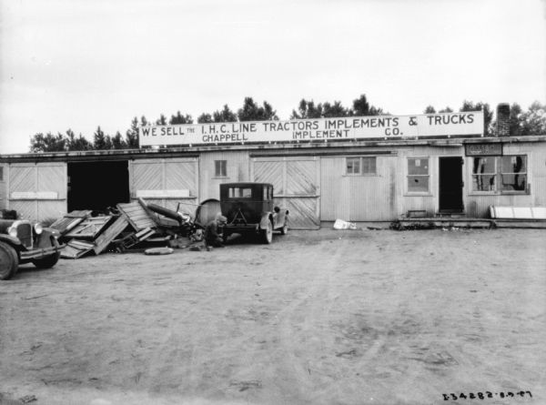 View across yard towards a man working at the rear of a car. The car is parked in front of a closed garage door of a building, with a sign that reads: "We Sell the I.H.C. Line Tractors Implements & Trucks, Chappell Implement Co."