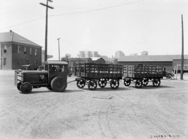 View across yard towards a man driving an industrial tractor pulling two stake-sided wagons. Industrial buildings are in the background.