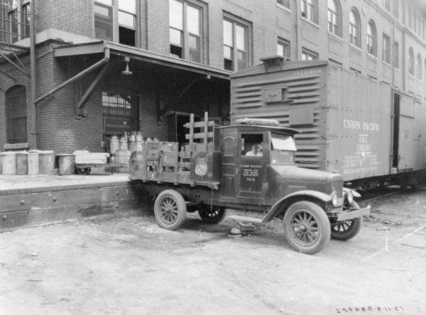 View of a man sitting in the driver's seat of a delivery truck backed up to a loading dock. The sign painted on the passenger door reads: "Union Pacific, Dining car and Hotel Department." Behind the truck is a railroad car along the loading dock.