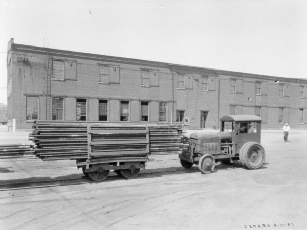 Right side profile view of a man driving an industrial tractor pushing a cart in front of it along railroad tracks. There is an industrial building in the background.