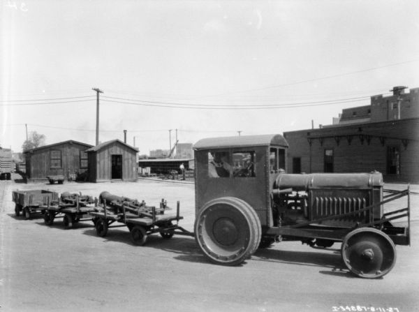 A man is driving an industrial tractor pulling three carts behind it. There are parts resting on the open carts. Industrial buildings are in the background.