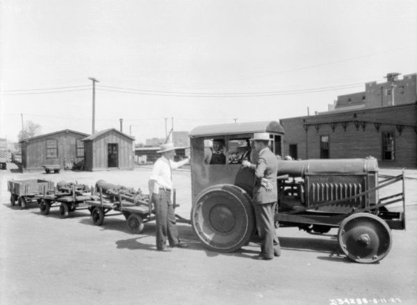 Two men are standing next to a man sitting in the driver's seat of an industrial tractor. The tractor is adapted to run on railroad tracks, and three carts are hitched to the back of the tractor.