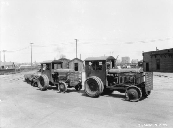 Two men are driving industrial tractors. The tractor on the left is pulling carts. In the background are industrial buildings and railroad tracks.