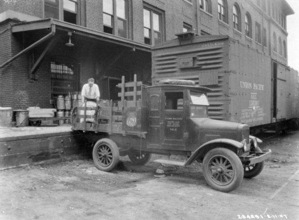 View of a man standing on a loading dock behind a delivery truck backed up to the loading dock. The sign painted on the passenger door reads: "Union Pacific, Dining car and Hotel Department." Behind the truck is a railroad car along the loading dock.