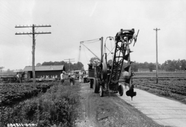 View towards a truck with equipment for installing telephone poles. There is a large auger suspended from the back of the truck. Men are standing along the side of the road on the left.
