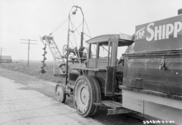 View towards front of truck parked along a road, with an auger hanging off the front. A man is standing, (obscured) at the back of the truck.