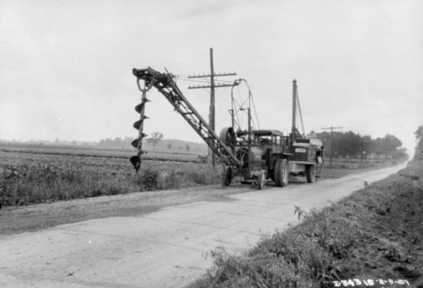 View across road towards a truck parked on the side of the road. A large auger is hanging in front of the truck. Fields are in the background.