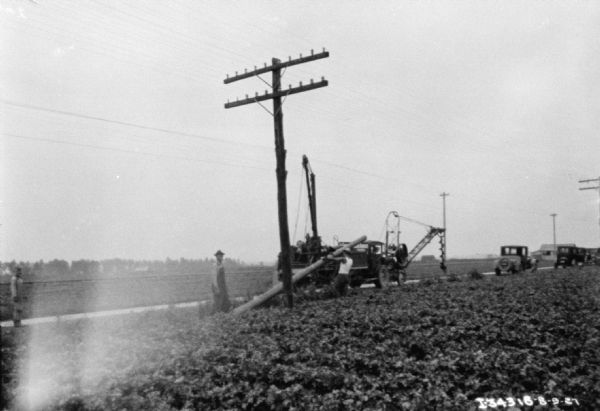 Men installing a telephone pole in a field. A truck is parked along a road behind them, and a large auger is attached to an apparatus at the front of the truck. A young child is standing in the road on the far left.