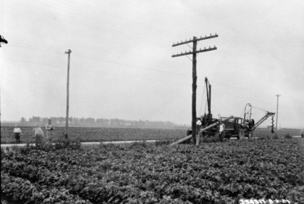 Men installing a telephone pole in a field. A truck is parked along a road behind them, and a large auger is attached to an apparatus at the front of the truck. Young children are standing in the road on the far left.
