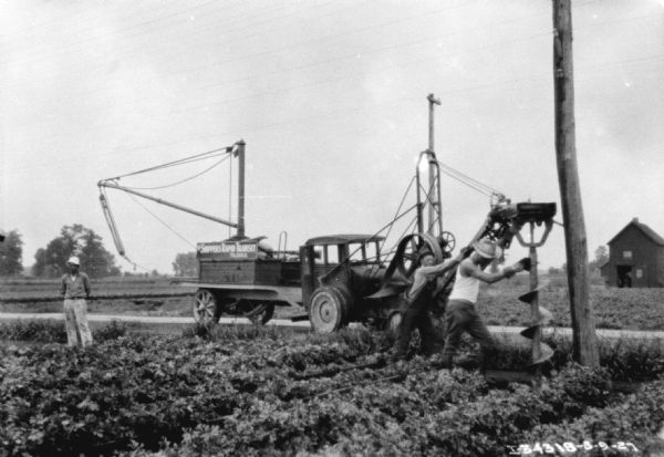 Two men are using a large auger to dig a hole for installing telephone poles. A truck is parked nearby, used for installing the poles. The sign on the side of the truck reads: "Shippers Rapid Transit, Toledo, O." Another man is standing in the field on the far left.