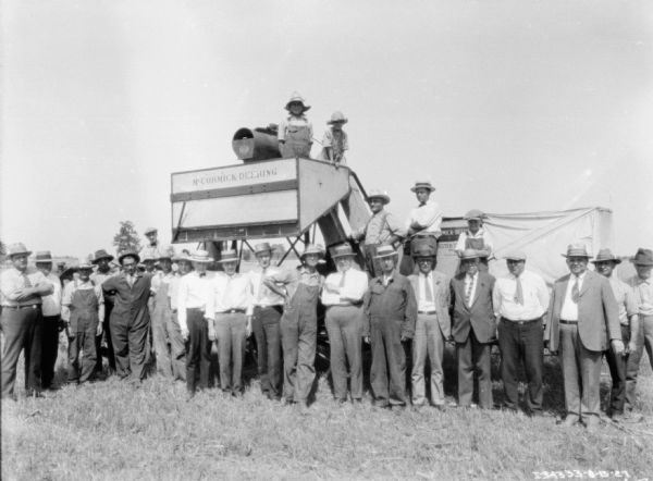 Group portrait of men standing in front of a McCormick-Deering harvester thresher in a field. Two children are standing on the harvester thresher above the group.