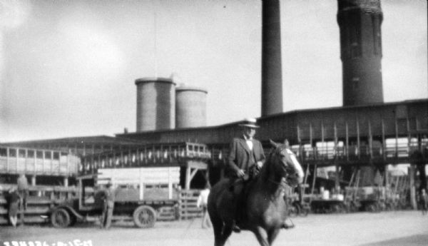A man is on horseback at a fair. Behind him are trucks along a fence, and large buildings and smokestacks.